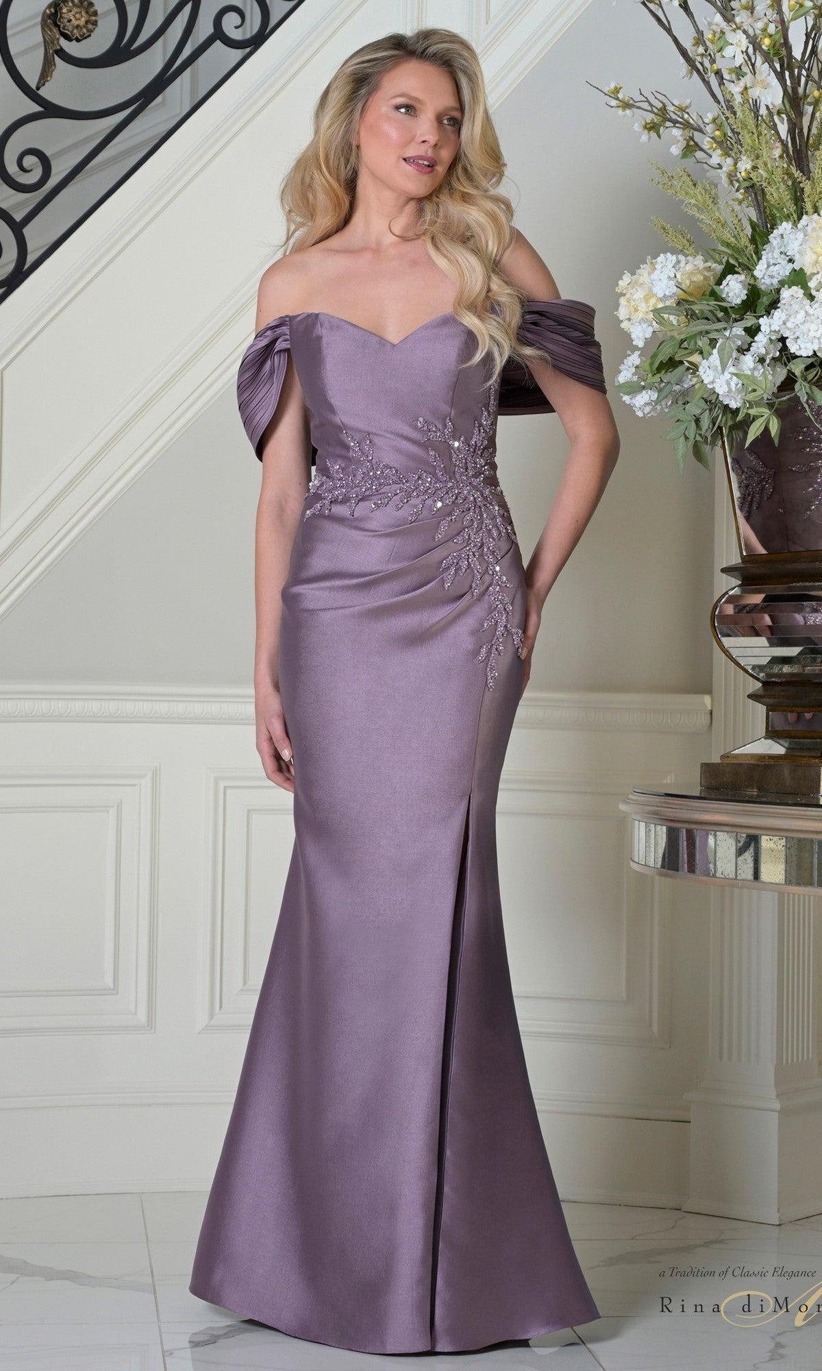 A woman stands indoors wearing the Rina Di Montella RD3527 long formal dress in Victorian, an off-the-shoulder, floor-length mermaid style with floral embroidery and draped sleeves; a staircase and flowers appear in the background.